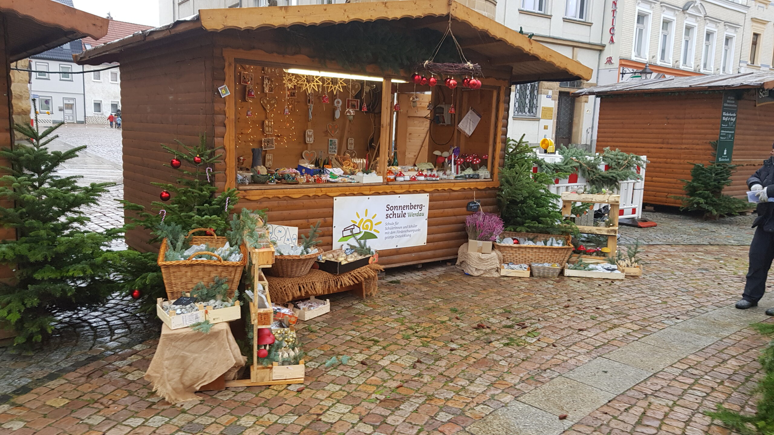 Der Stand der Sonnenbergschule auf dem Weihnachtsmarkt in Werdau. Auf dem Bild sieht man eine Weihnachtsmarktbude aus Holz, An der Frontseite der Bude ist das Logo der Schule zu sehen. Rechts und Links neben der Bude stehen noch einige Artikel, welche dort ebenfalls verkauft werden.