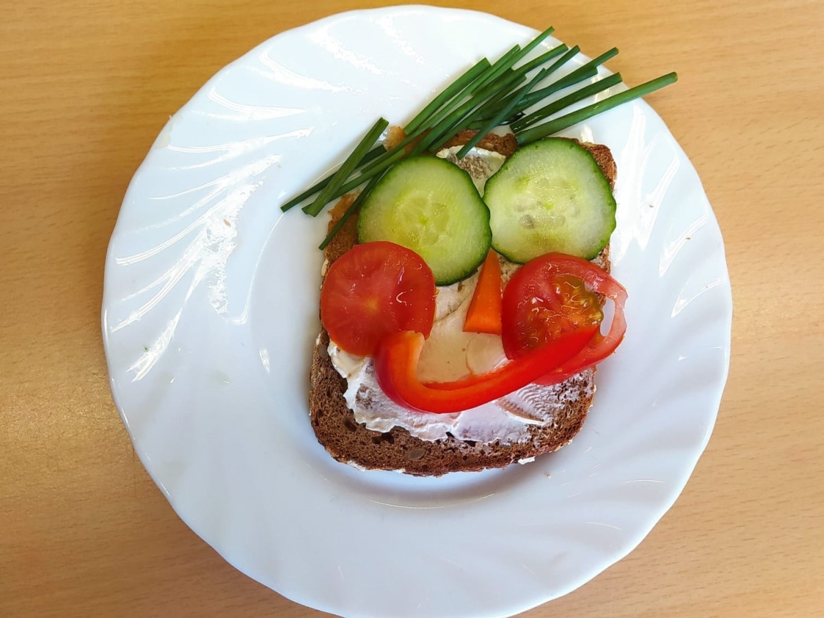 Eine Scheibe Kastenbrot mit zwei Gurkenscheiben als Augen, Tomatenscheiben als Wangen, einem Dreieck aus Paprika als Nase und einem langen, gebogenen Paprikastück als Mund.