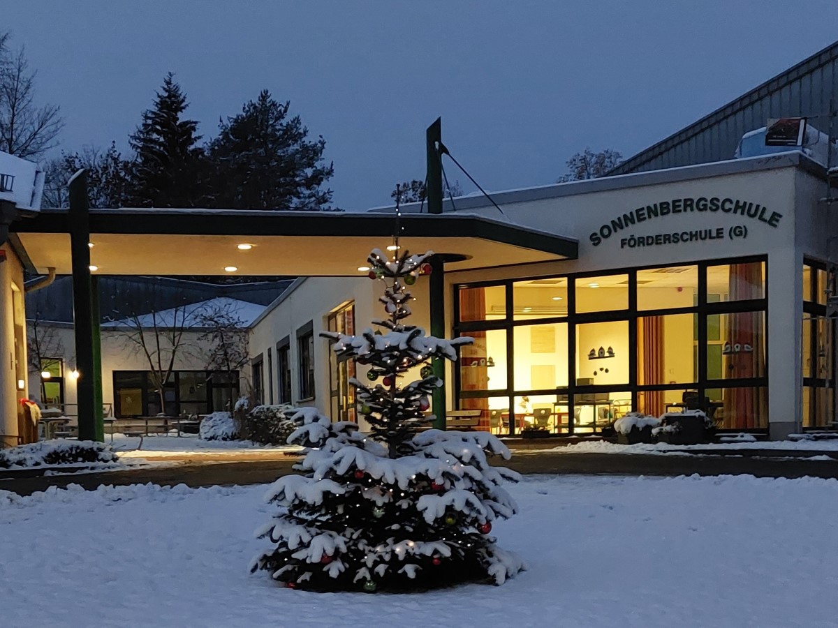 Ein Bild von der eingeschneiten Schule. Im Vordergrund steht eine kleine Tanne, welche mit einer Lichterkette geschmückt ist.